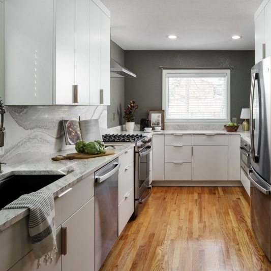 An interior view of a recently remodeled KCMO kitchen featuring sleek white cabinets, stainless steel appliances, a gray accent wall, and a light wood floor.