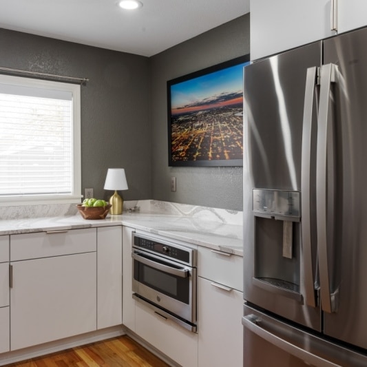 An interior view of a recently remodeled KCMO kitchen featuring sleek white cabinets, stainless steel appliances, a gray accent wall with a cityscape picture, and a light wood floor.