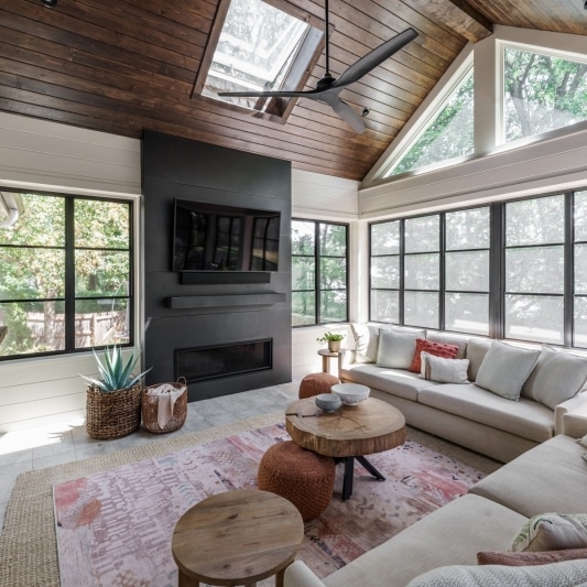 newly added porch addition on back of home with warm dark wood slats on ceiling and white creamy walls with black framed windows and a dark charcoal colored statement media wall fireplace feature