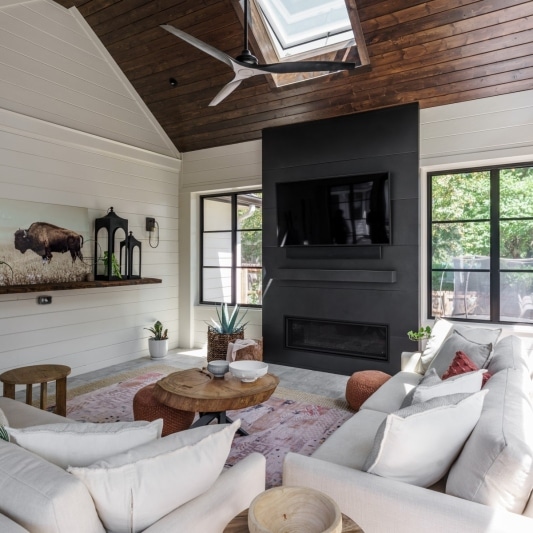 newly added porch addition on back of home with warm dark wood slats on ceiling and white creamy walls with black framed windows and a dark charcoal colored statement media wall fireplace feature