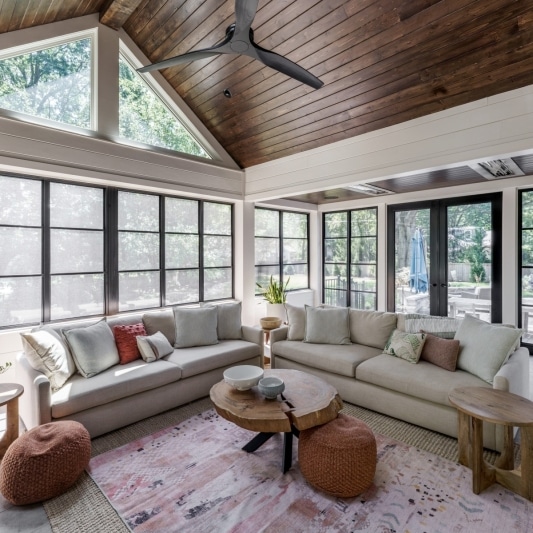 newly added porch addition on back of home with warm dark wood slats on ceiling and white creamy walls with black framed windows and a dark charcoal colored statement media wall fireplace feature