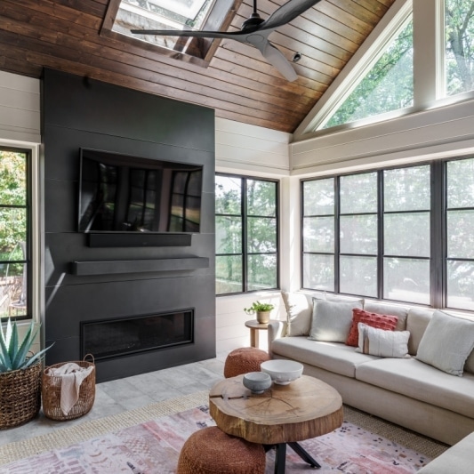 newly added porch addition on back of home with warm dark wood slats on ceiling and white creamy walls with black framed windows and a dark charcoal colored statement media wall fireplace feature