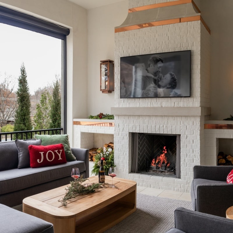Cozy outdoor lanai with a white brick wood-burning fireplace, copper accents, built-in firewood storage, and modern seating area.