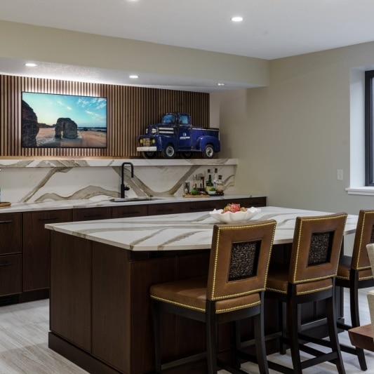 Modern basement bar with a large island, marble-look countertops, and a mounted TV.
