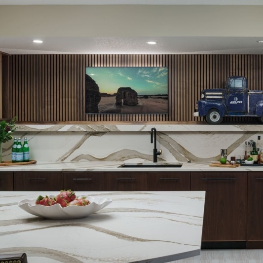 Modern basement bar with a large island, marble-look countertops, and a mounted TV.