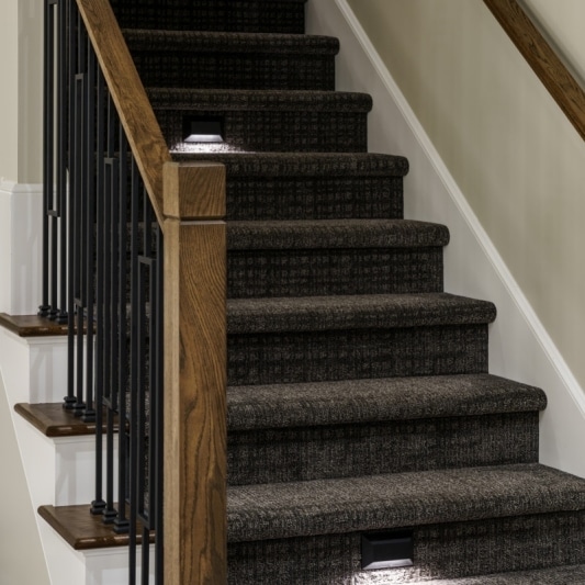 Modern basement stairs with dark carpet, wood banister, metal balusters, and built-in step lights.