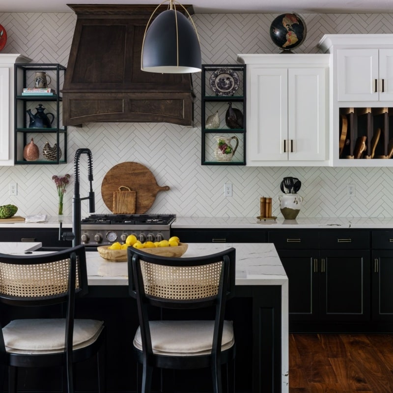 An interior view of a stylish kitchen featuring a Mont Blanc quartzite island with a white countertop, black lower cabinets, white upper cabinets, a dark wood range hood, and herringbone backsplash.