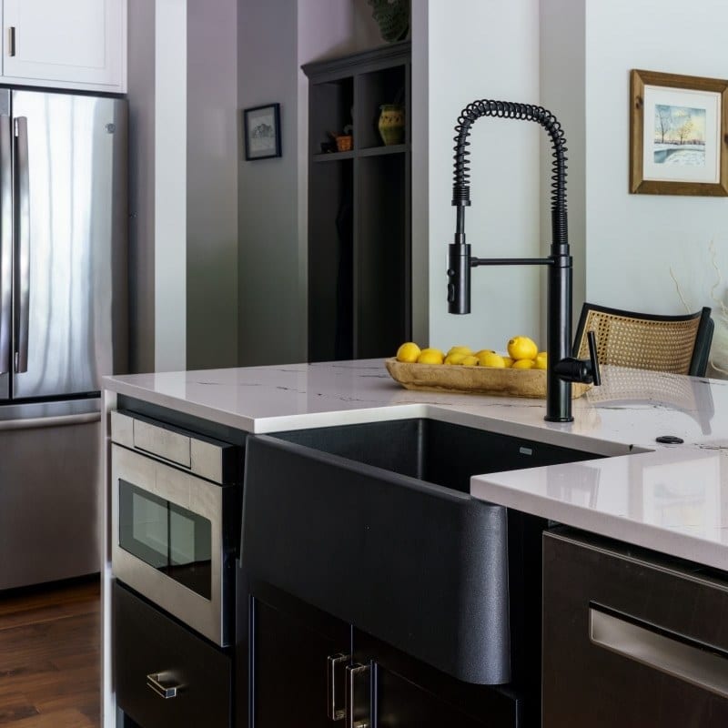 Interior view of a well-lit kitchen featuring a U-shaped layout with warm wood grain cabinets, a multi-colored backsplash, a white countertop, and a central gray kitchen island with a contrasting white oval countertop. Two geometric black pendant lights hang above the island.