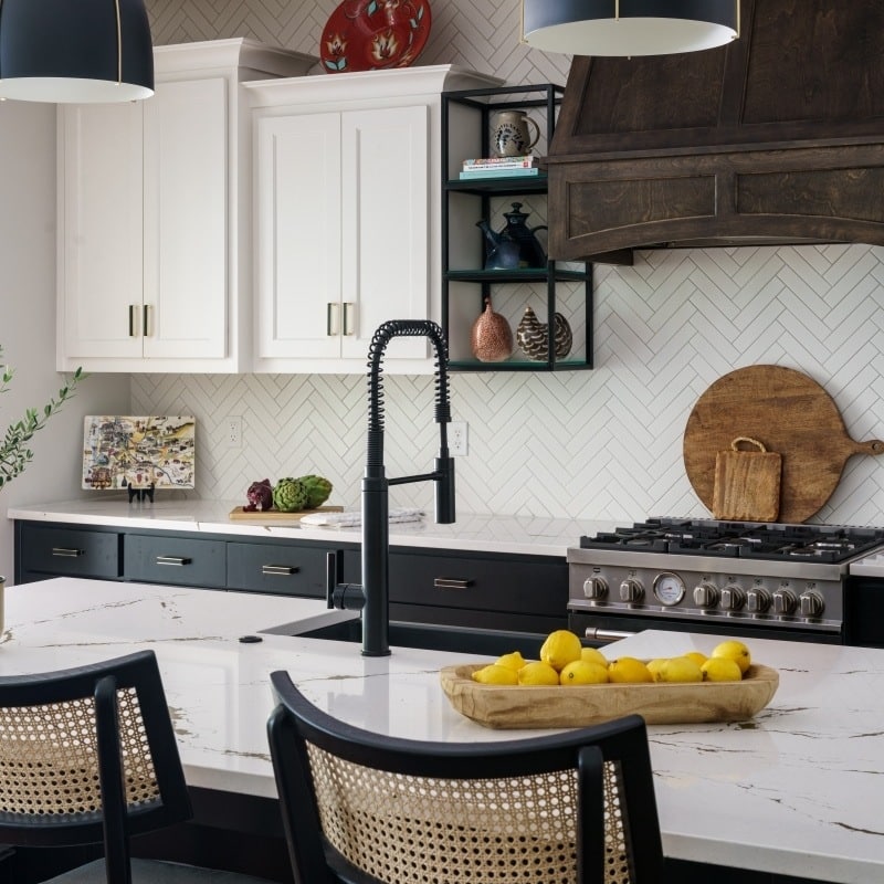 An interior view of a stylish kitchen featuring a Mont Blanc quartzite island with a white countertop, black lower cabinets, white upper cabinets, a dark wood range hood, and herringbone backsplash.