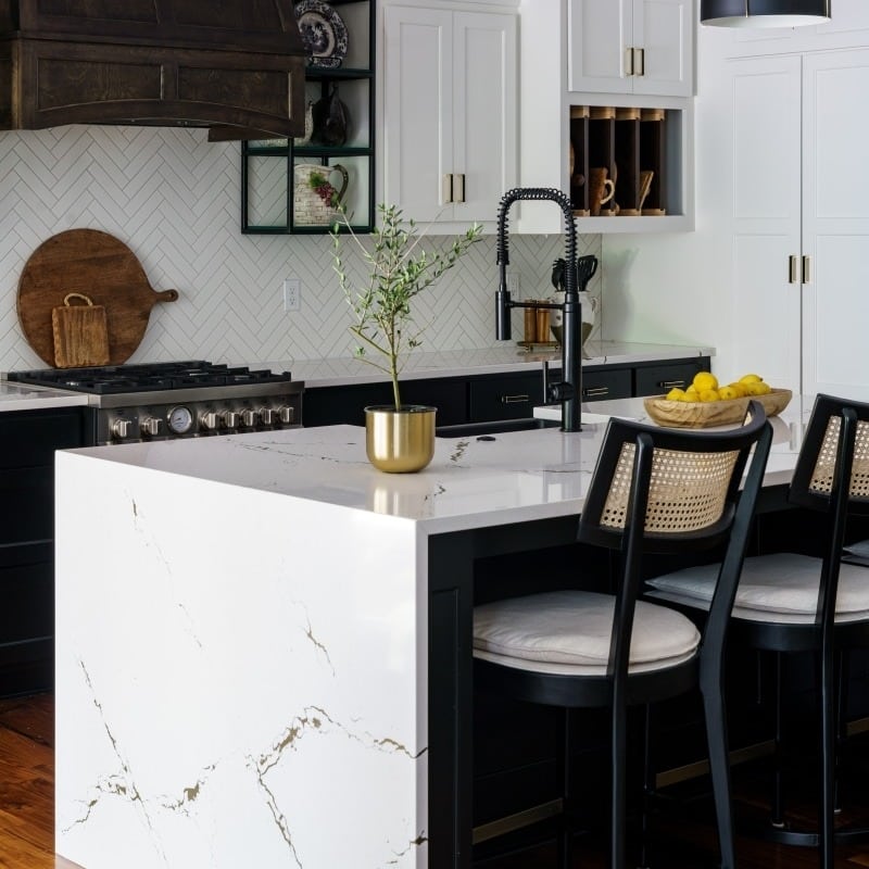 An interior view of a stylish kitchen featuring a Calacatta Novus island with a white countertop, black lower cabinets, white upper cabinets, a dark wood range hood, and herringbone backsplash.