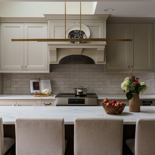 A recently remodeled kitchen featuring light gray shaker-style cabinets, a large white marble-look island with seating for four, stainless steel appliances, a light beige subway tile backsplash, and a unique linear brass pendant light fixture above the island.