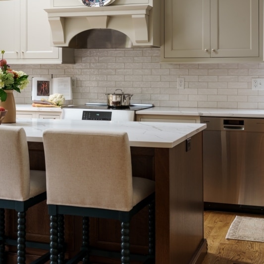 A recently remodeled kitchen featuring light gray cabinets, a large white marble-look island with seating, stainless steel appliances, a light beige subway tile backsplash, and a unique linear brass pendant light fixture above the island.