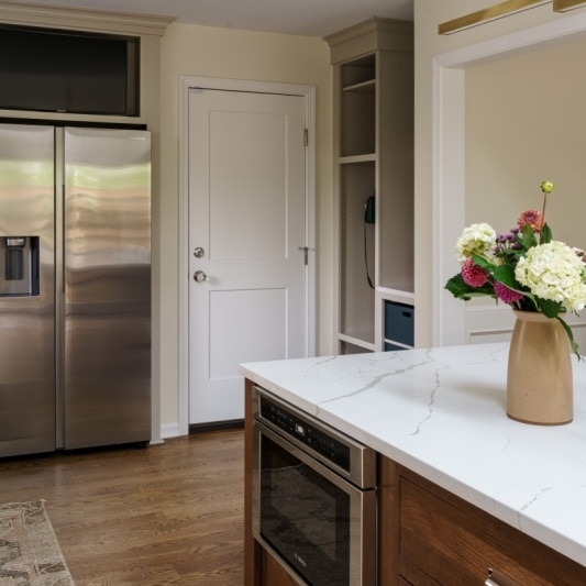 A kitchen with two-toned cabinets, featuring a dark wood island with a white countertop and a stainless steel microwave, and stainless steel refrigerator.