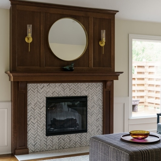 A living room featuring a fireplace with a herringbone tile surround, a dark wood mantel, and a round mirror.