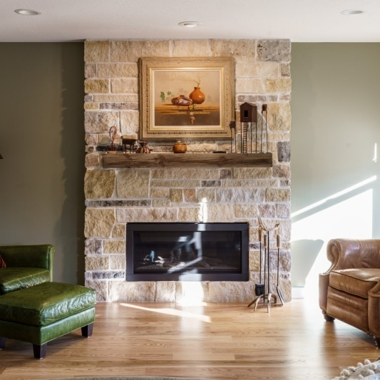 An interior view of a living room featuring a stone fireplace, a brown leather recliner, a green armchair with an ottoman, and a wooden floor.