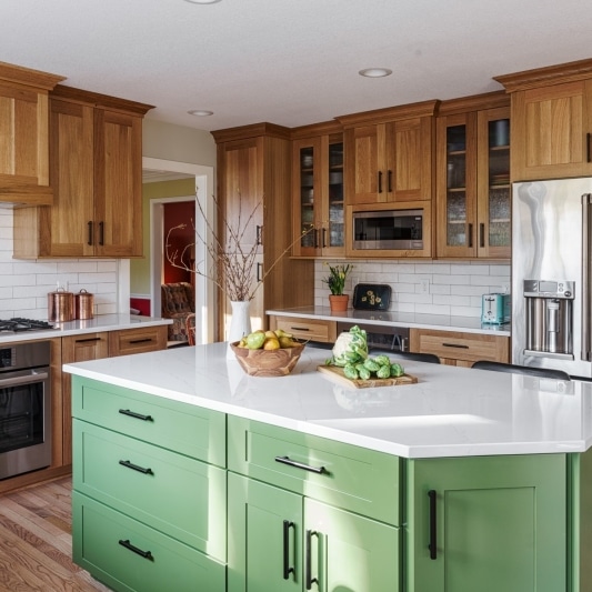 An interior view of a well-lit kitchen featuring light wood cabinets, a green kitchen island with a white countertop, and stainless steel appliances.