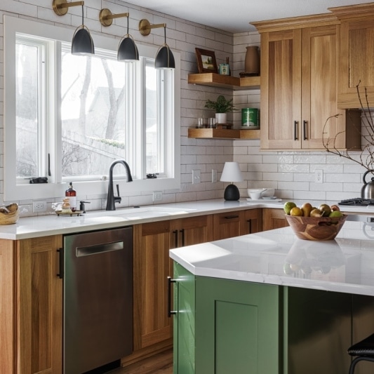An interior view of a well-lit kitchen featuring warm wood cabinetry, a green kitchen island with a white countertop, and three black pendant lights above the sink.