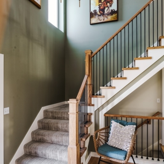 An interior view of a staircase featuring gray walls, a wooden railing with black spindles, a woven chair, and colorful artwork.