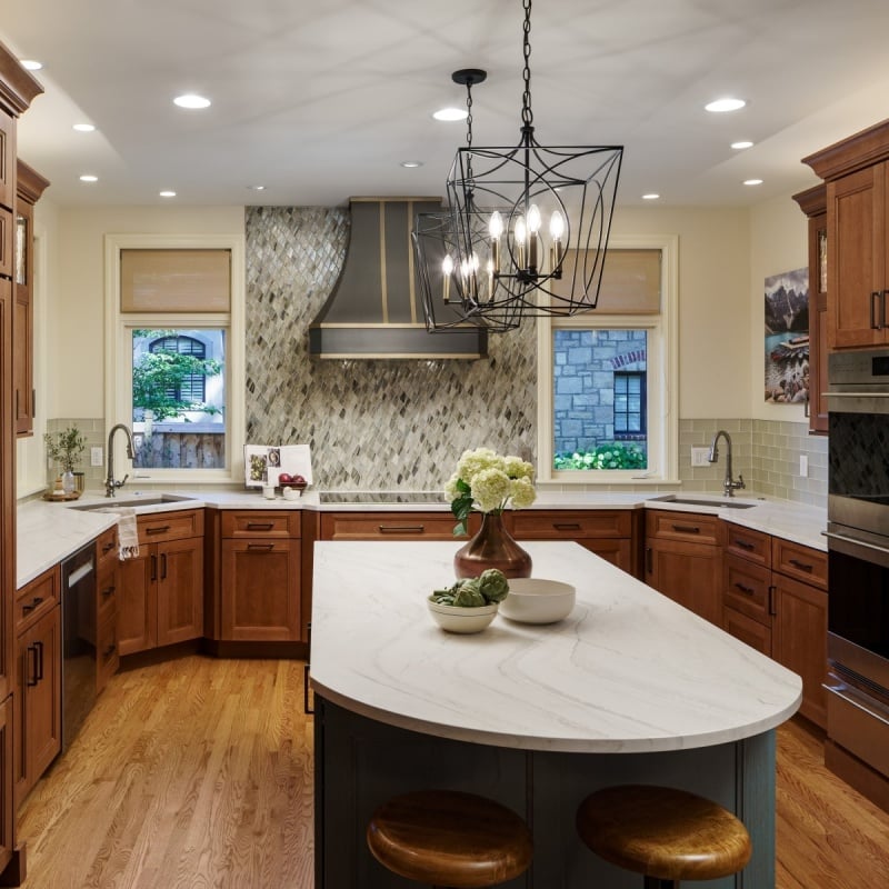 A beautifully remodeled kitchen in Mission Hills, Kansas, featuring warm wood cabinets, a large curved white countertop island with seating, a decorative range hood, and modern pendant lighting.