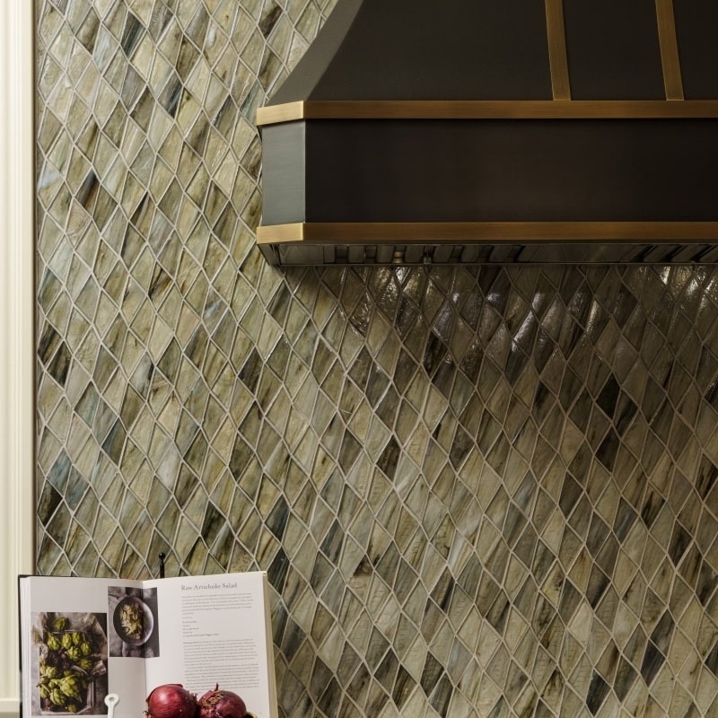 A detailed view of a kitchen showcasing a textured diamond-patterned backsplash in muted green and gray tones, a dark metal range hood with brass accents, and a white countertop with various kitchen items.