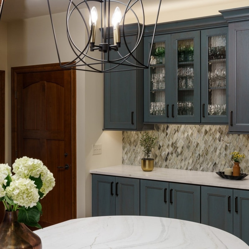 Close-up of a well-lit kitchen storage area featuring blue-gray cabinets with glass doors, displaying glassware and bottles, with a patterned backsplash and a portion of a round white countertop.