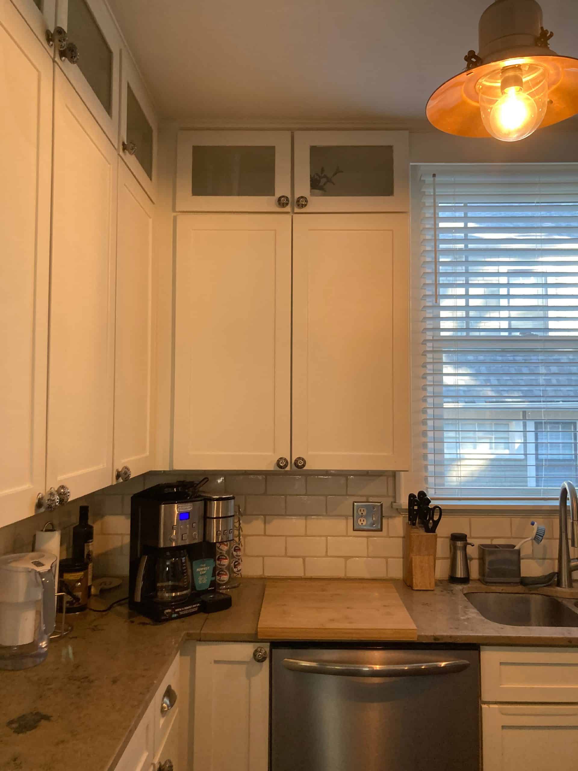 Before image of a crammed and dated kitchen with white cabinets and basic white tile backsplash similar to subway tile and grey dark counters