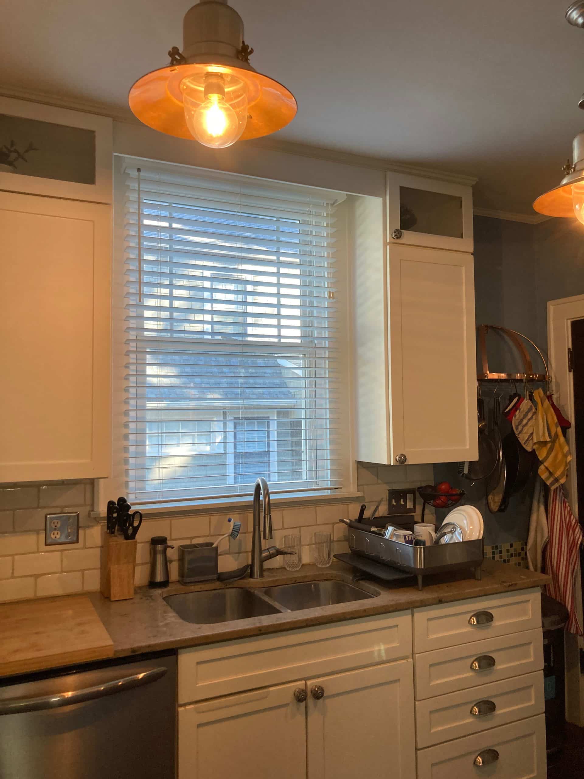 before photo of the kitchen sink and window and white cabinets. there is a sort of cluttered and tight feeling when you look at the space