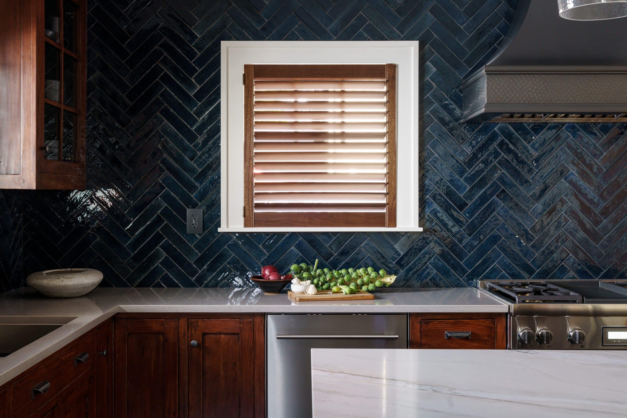 This image showcases a refreshed kitchen detail, focusing on a window area. The window, framed in white, features light brown wooden slatted blinds. Below the window, a deep blue herringbone tile backsplash adds a vibrant touch. Warm-toned wooden cabinetry is present both below the counter and above, with the upper cabinets featuring glass doors displaying glassware. A silver faucet is visible against the backsplash. On the counter, a few pieces of produce are arranged. To the left, a glimpse of a plant with a large orange flower adds a natural element. The overall style blends classic and contemporary elements, creating a cozy yet stylish kitchen space.