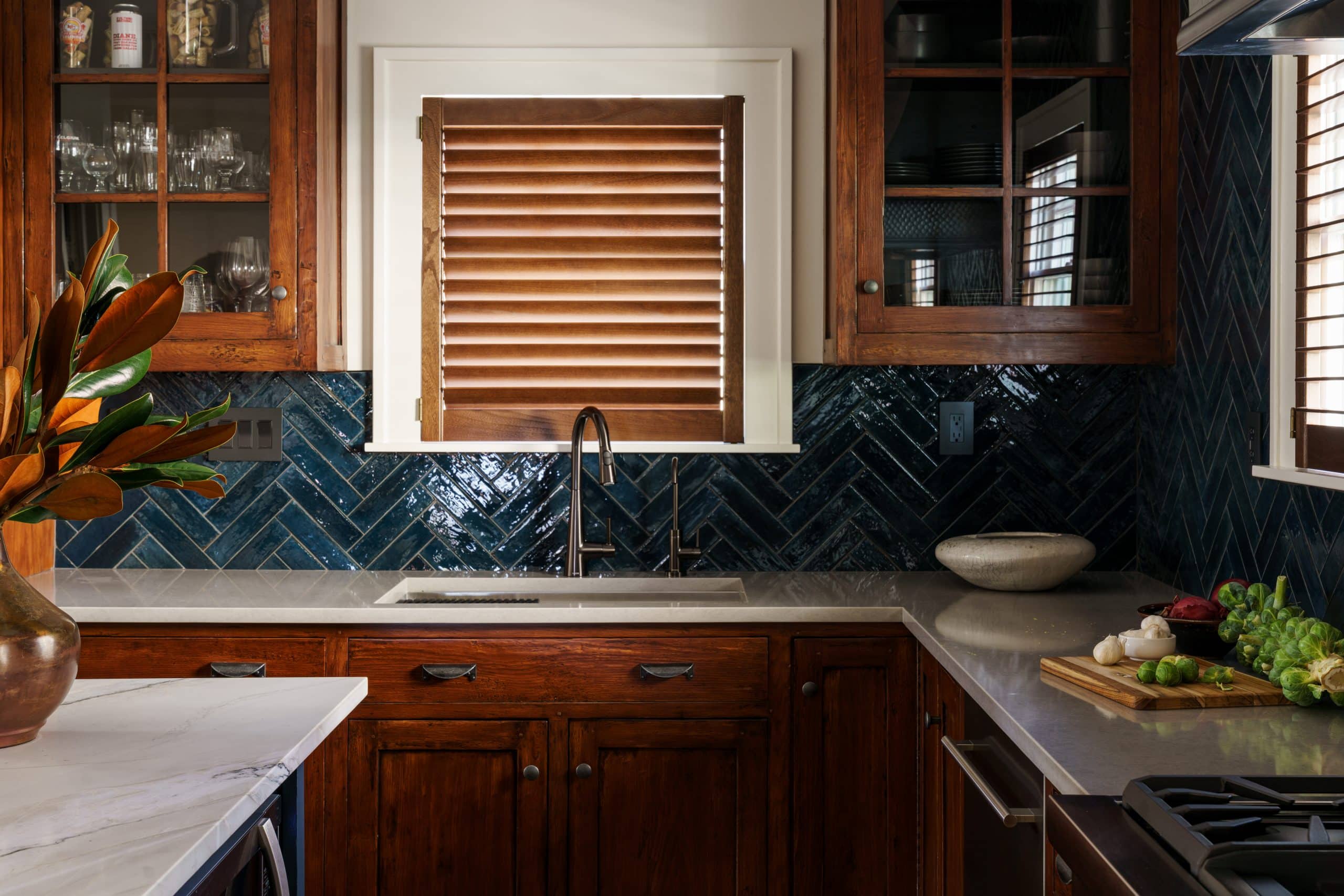 This image showcases a refreshed kitchen detail, focusing on a window area. The window, framed in white, features light brown wooden slatted blinds. Below the window, a deep blue herringbone tile backsplash adds a vibrant touch. Warm-toned wooden cabinetry is present both below the counter and above, with the upper cabinets featuring glass doors displaying glassware. A silver faucet and a stainless steel sink are visible to the left, set into the light countertop. On the counter, a few pieces of produce are arranged. The overall style blends classic and contemporary elements, creating a cozy yet stylish kitchen space.