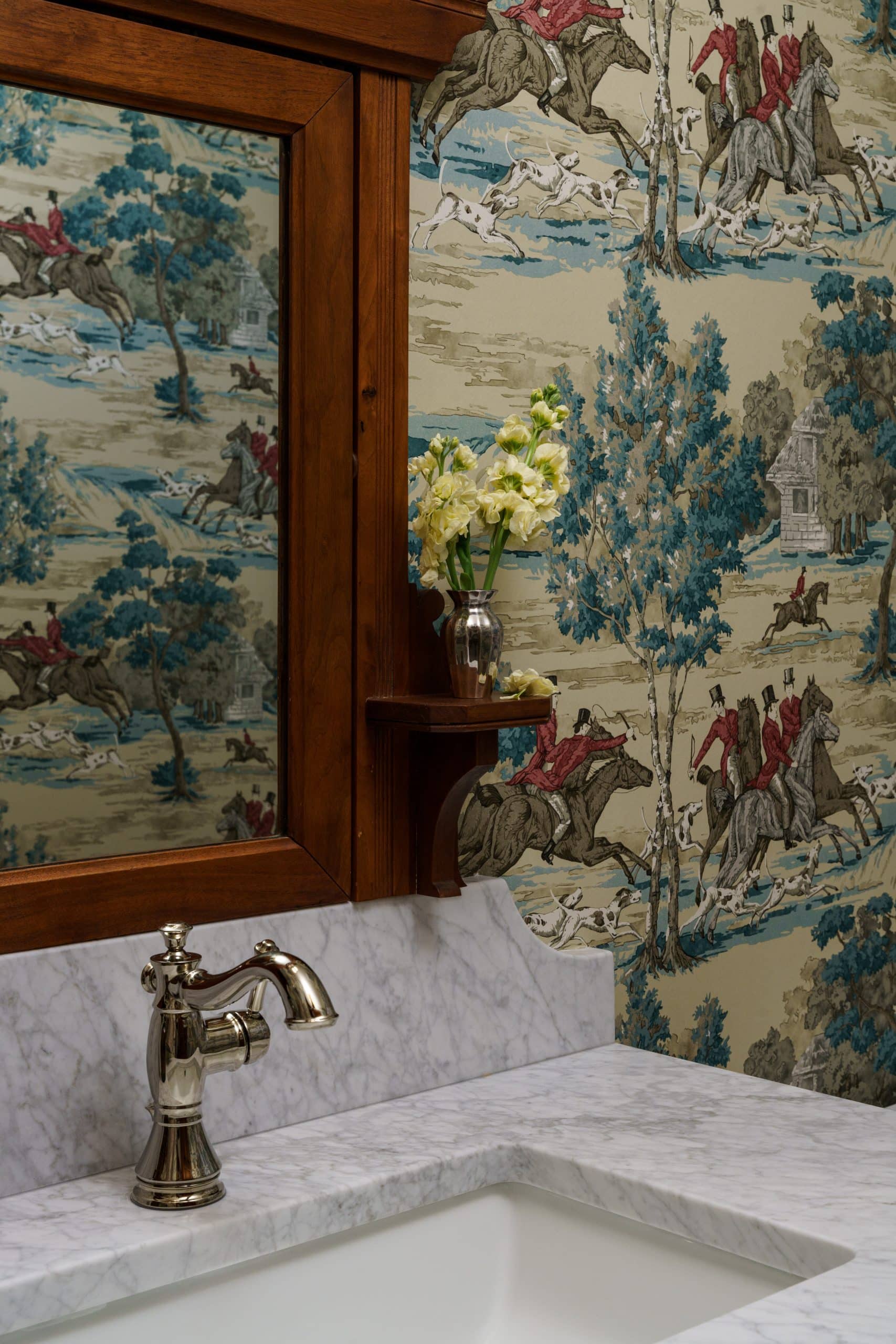 Close-up detail of a charming powder bathroom, showcasing a white marble countertop and backsplash, an undermount sink, and a classic polished nickel faucet. In the background, a portion of the family heirloom vintage wood mirror with a small shelf is visible, reflecting the whimsical fox hunt pattern of the wallpaper in blue, red, and beige tones