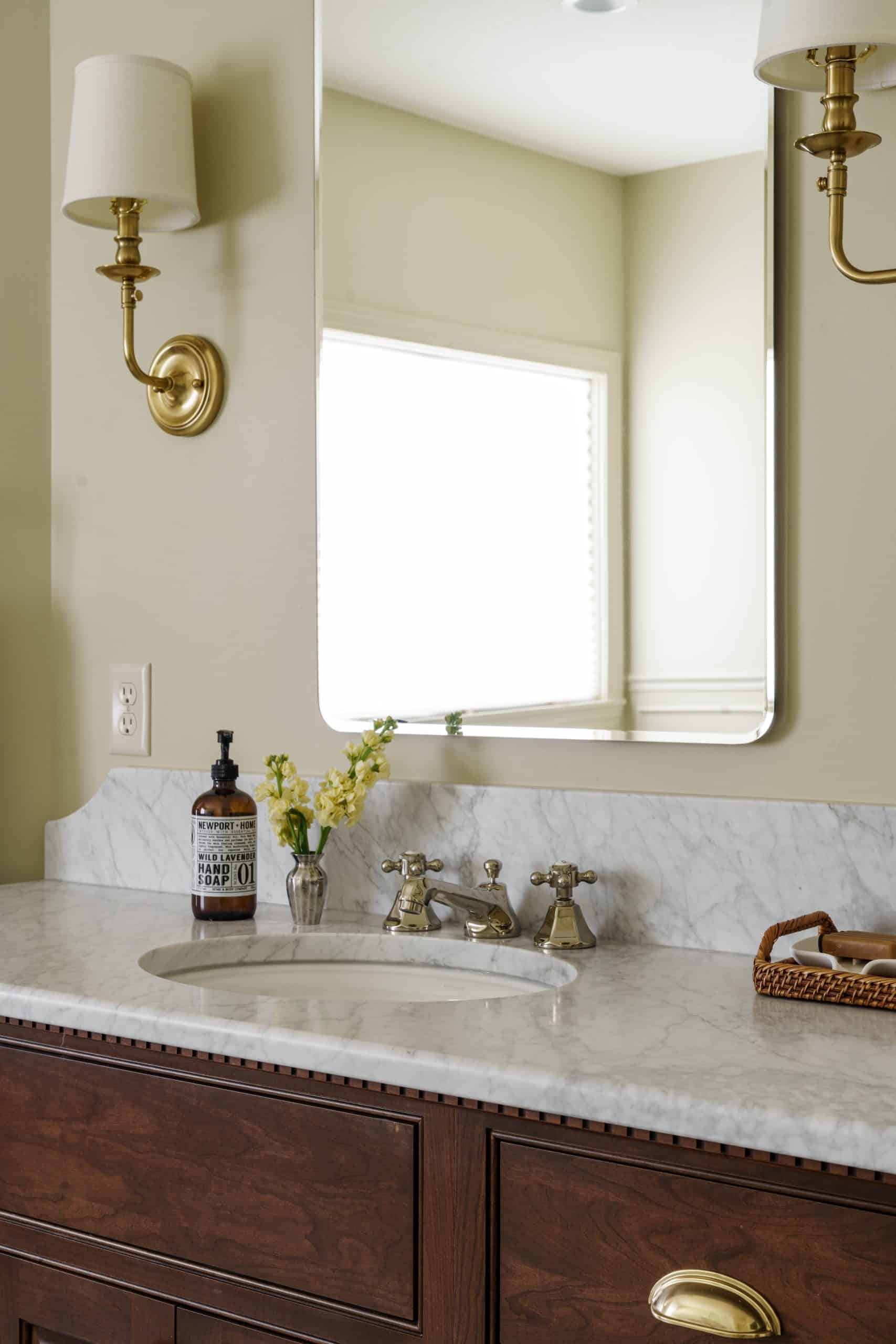 Close-up of one sink area on a remodeled primary bathroom vanity, showing a white undermount sink set into a white marble countertop with a sculpted backsplash. A classic polished nickel or chrome faucet, a decorative soap dispenser, and small flowers are visible. Above, a rectangular mirror reflects a window, flanked by an elegant brass sconce.