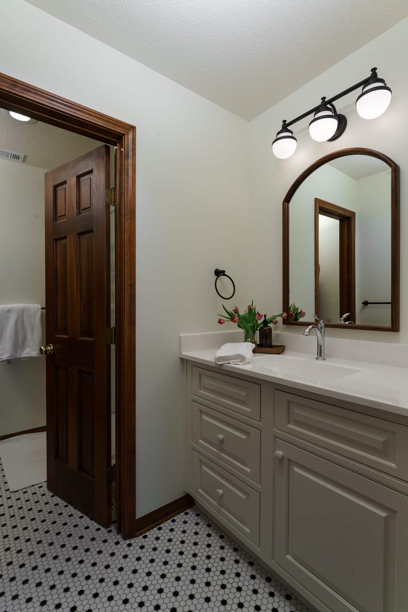 Son's side of a Jack and Jill bathroom, featuring a light-colored vanity with an undermount sink and a white countertop. Above the vanity, an arched wood-framed mirror is illuminated by a black three-light vanity fixture. The room has classic black and white hexagonal tile flooring and a dark wood door leading to the shared shower/toilet area.