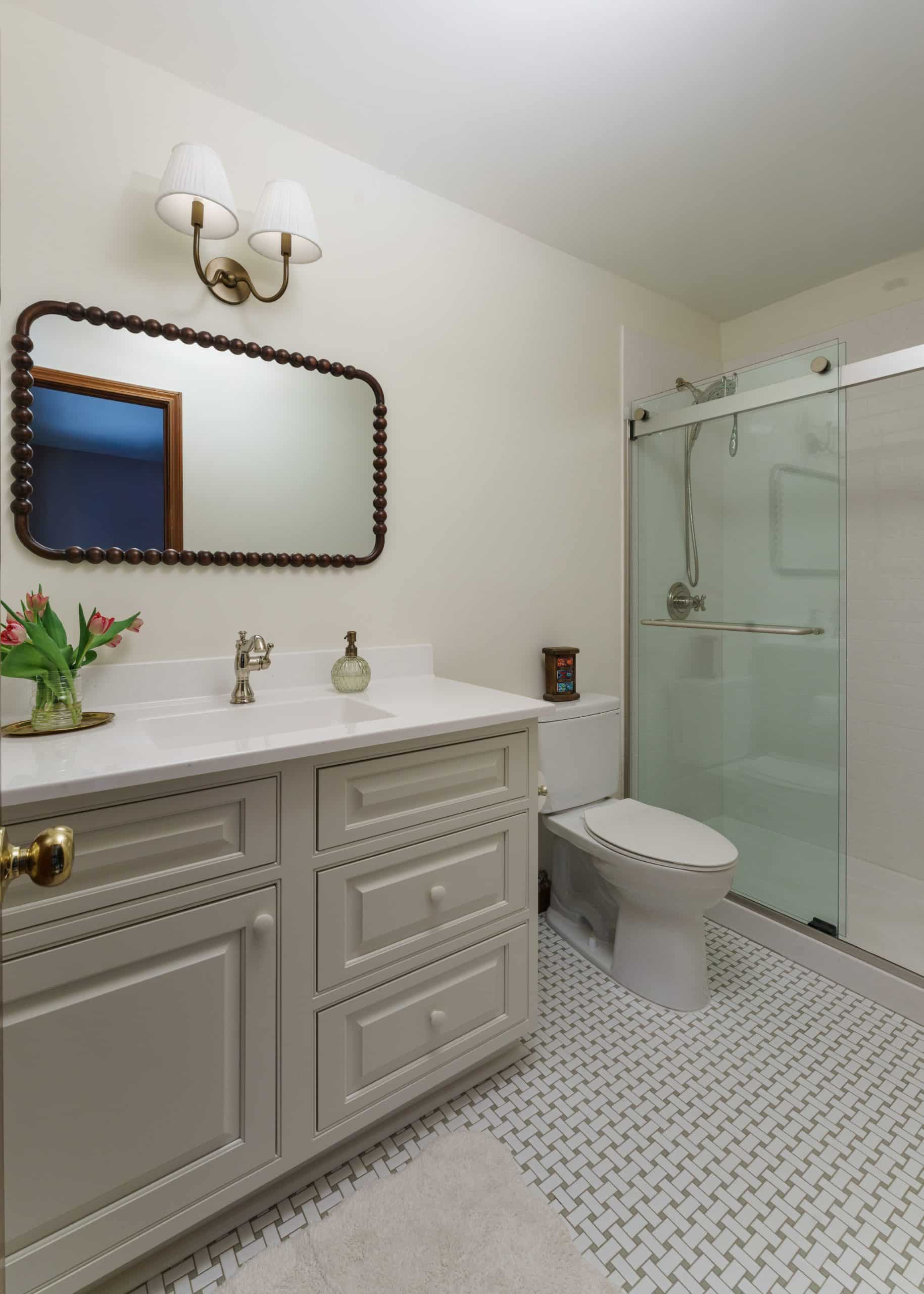 Light and classic daughter's bathroom, part of a Jack and Jill setup, featuring a white vanity with an undermount sink and light countertop, a unique rectangular mirror with a beaded dark wood frame, and elegant wall sconces. The room includes a modern toilet and a glass-enclosed shower, all set on a light-colored mosaic tile floor