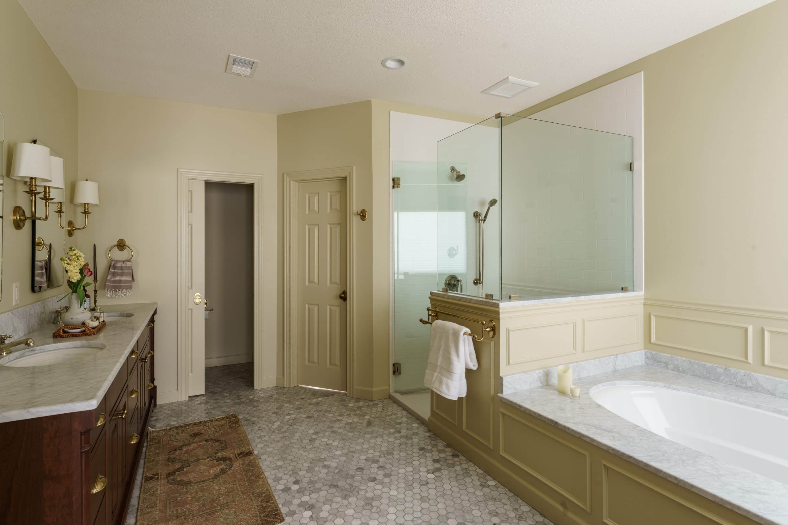 "A classic and charming primary bathroom remodel, showcasing the full scope of the luxurious design with a wainscoted tub, glass shower, and rich wood double vanity. The room's light palette and traditional elements create a timeless retreat.