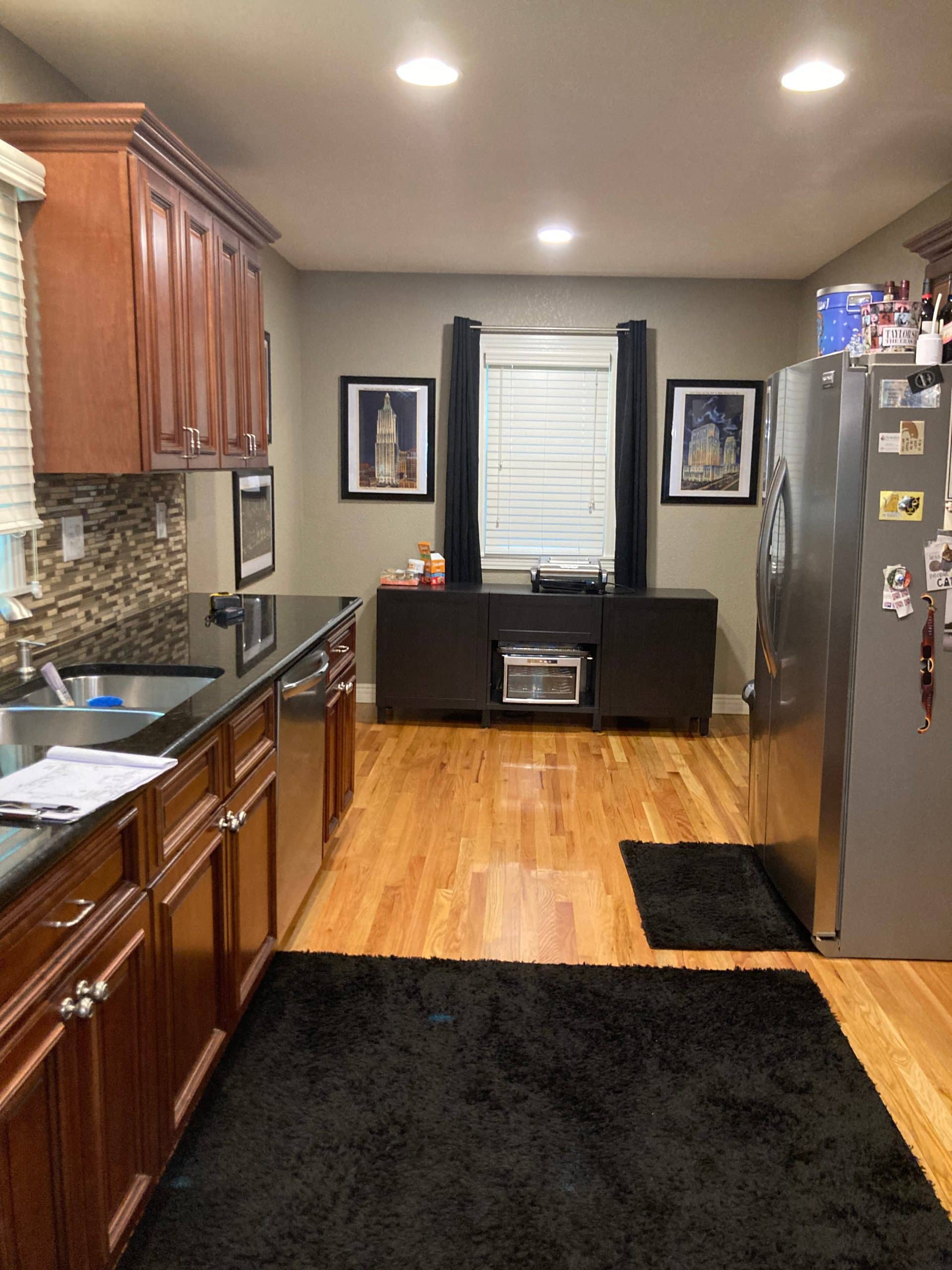 A wide shot of a dated kitchen featuring dark wood cabinets, black countertops, a mosaic tile backsplash, and light wood flooring. A large stainless steel refrigerator stands on the right, and a dark cabinet with a window above it is centered on the back wall. Black rugs are on the floor.