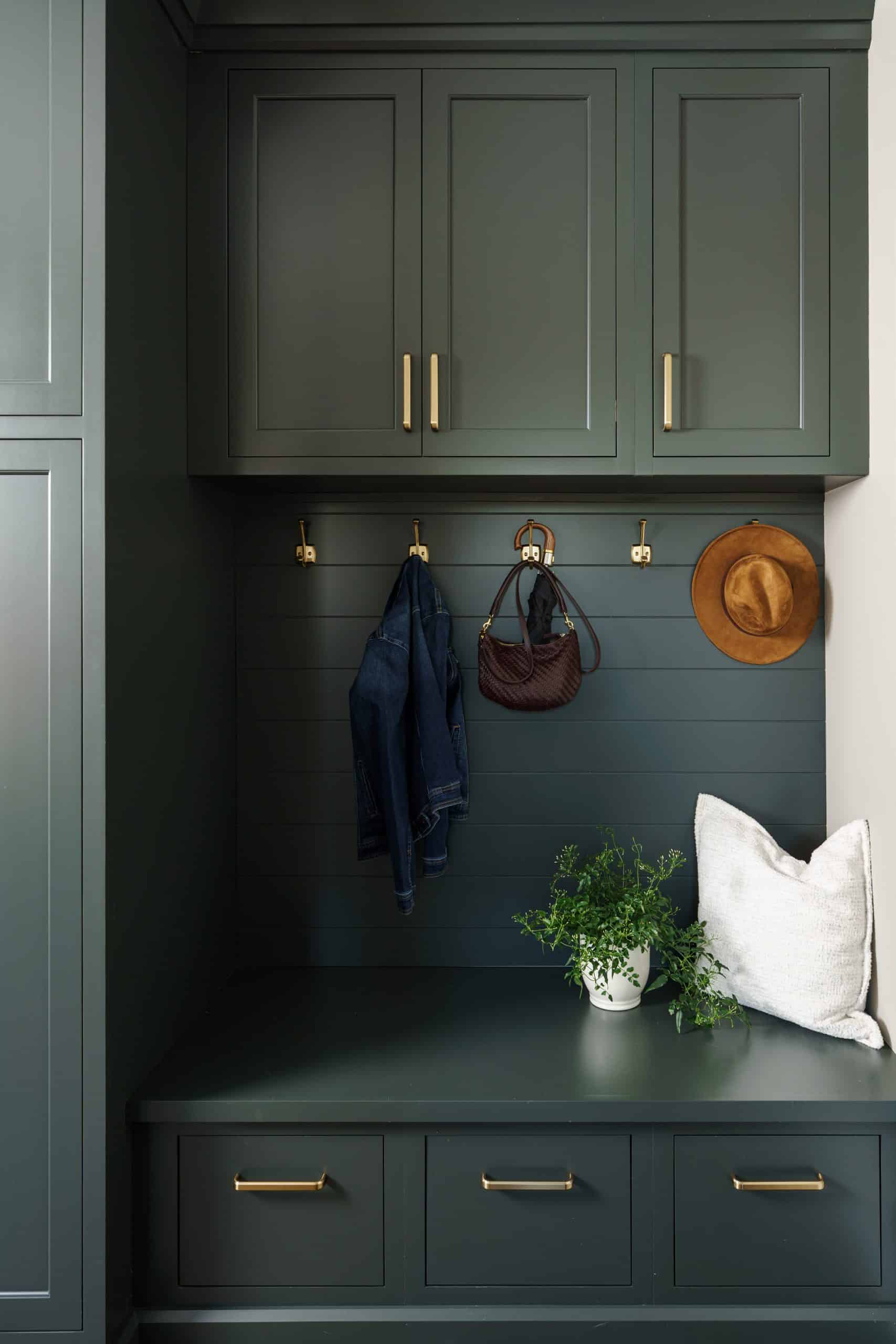 A stylish mudroom nook featuring dark gray-green cabinetry and a built-in bench seat with storage drawers. The wall behind the bench is finished with horizontal paneling in a matching color, with gold hooks holding a denim jacket, a handbag, and a wide-brimmed hat. A plush white pillow and a potted fern add a welcoming, homey touch to the space. This image highlights a functional and beautifully designed mudroom storage solution, perfect for a modern home