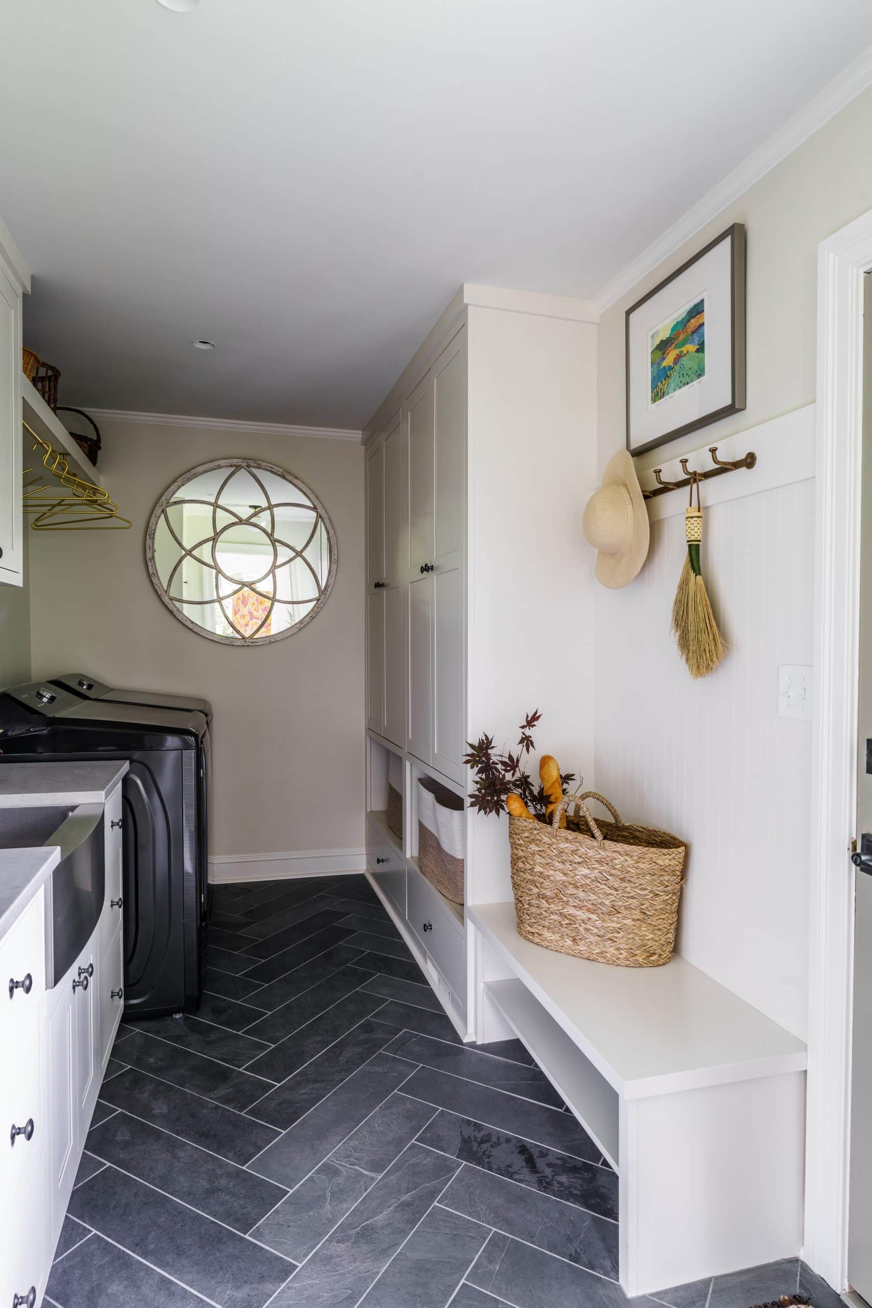 A bright and functional laundry room and mudroom combination. The space features white cabinetry and a large apron-front sink, with a washer and dryer on the left. The floor has dark gray herringbone-patterned tiles. A built-in bench with open storage below and a woven basket sits on the right. A decorative round mirror hangs on the wall, and a framed picture and a hat are on hooks above the bench. This image showcases a smart and stylish laundry and storage solution for a modern home.