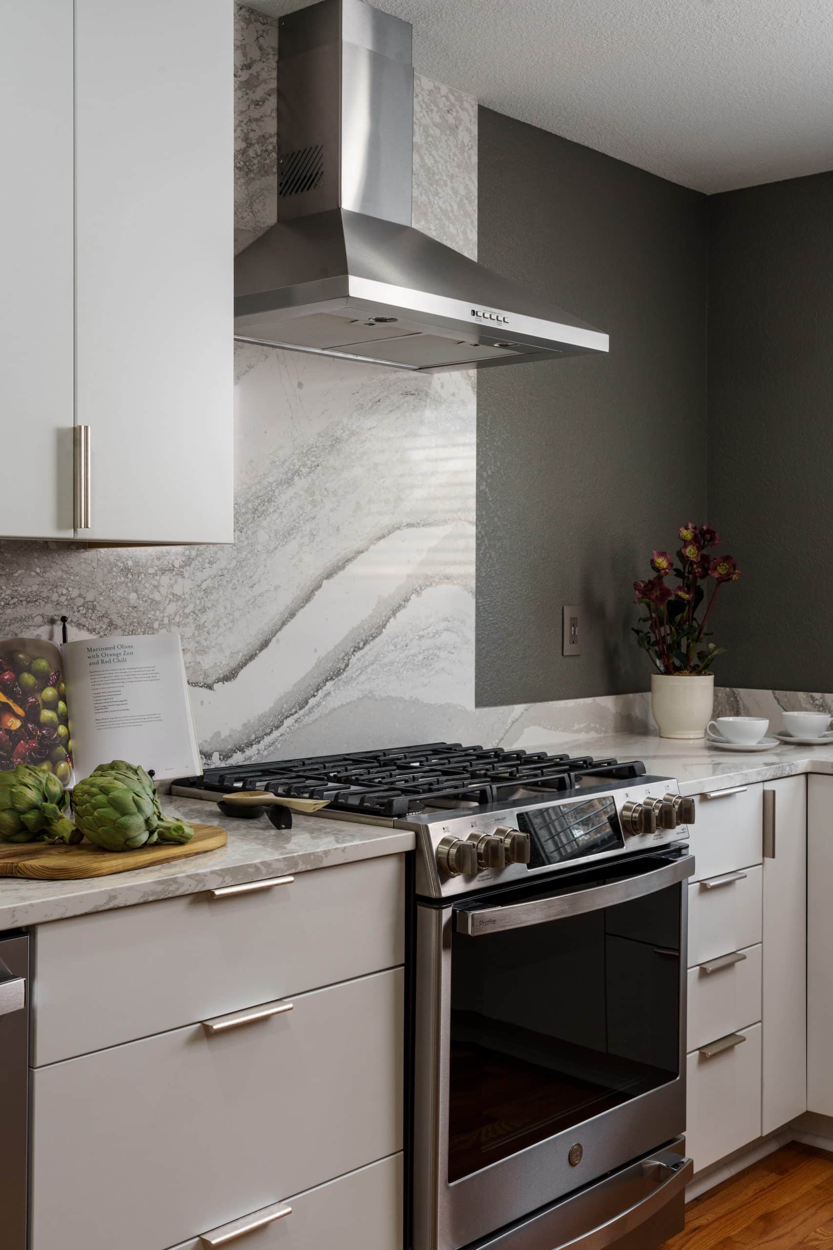 A close-up view of a modern kitchen featuring a stainless steel range and matching hood. The area behind the range has a full-height slab backsplash of light-colored, veined quartz. White flat-panel cabinets are on the left, and a dark gray wall is visible on the right.
