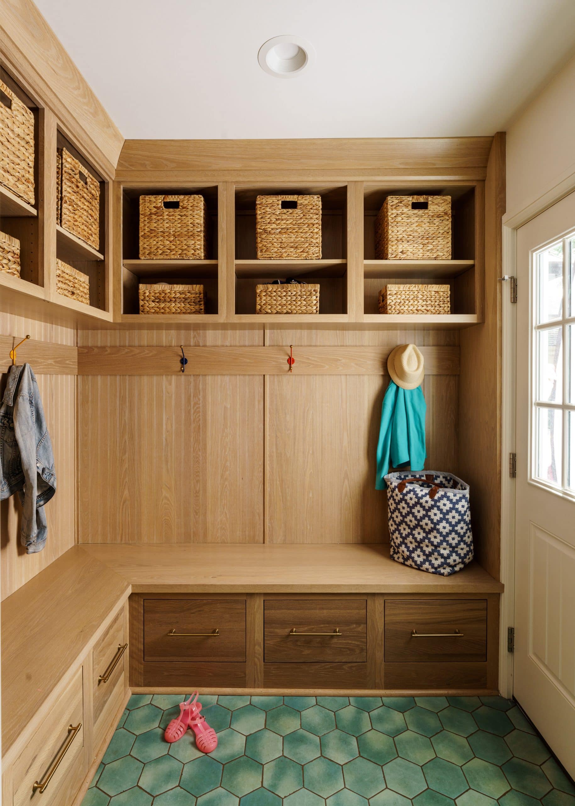 A modern mudroom featuring custom white oak cabinetry with open shelving and built-in benches. Woven baskets are organized in the upper shelves for a clean look. The space includes multiple hooks for hanging jackets and bags, and a built-in bench seat with large storage drawers below. The floor is tiled with stylish teal-colored hexagonal tiles, providing a pop of color and a durable surface. This image showcases a beautiful and functional white oak mudroom design, perfect for a family-friendly home