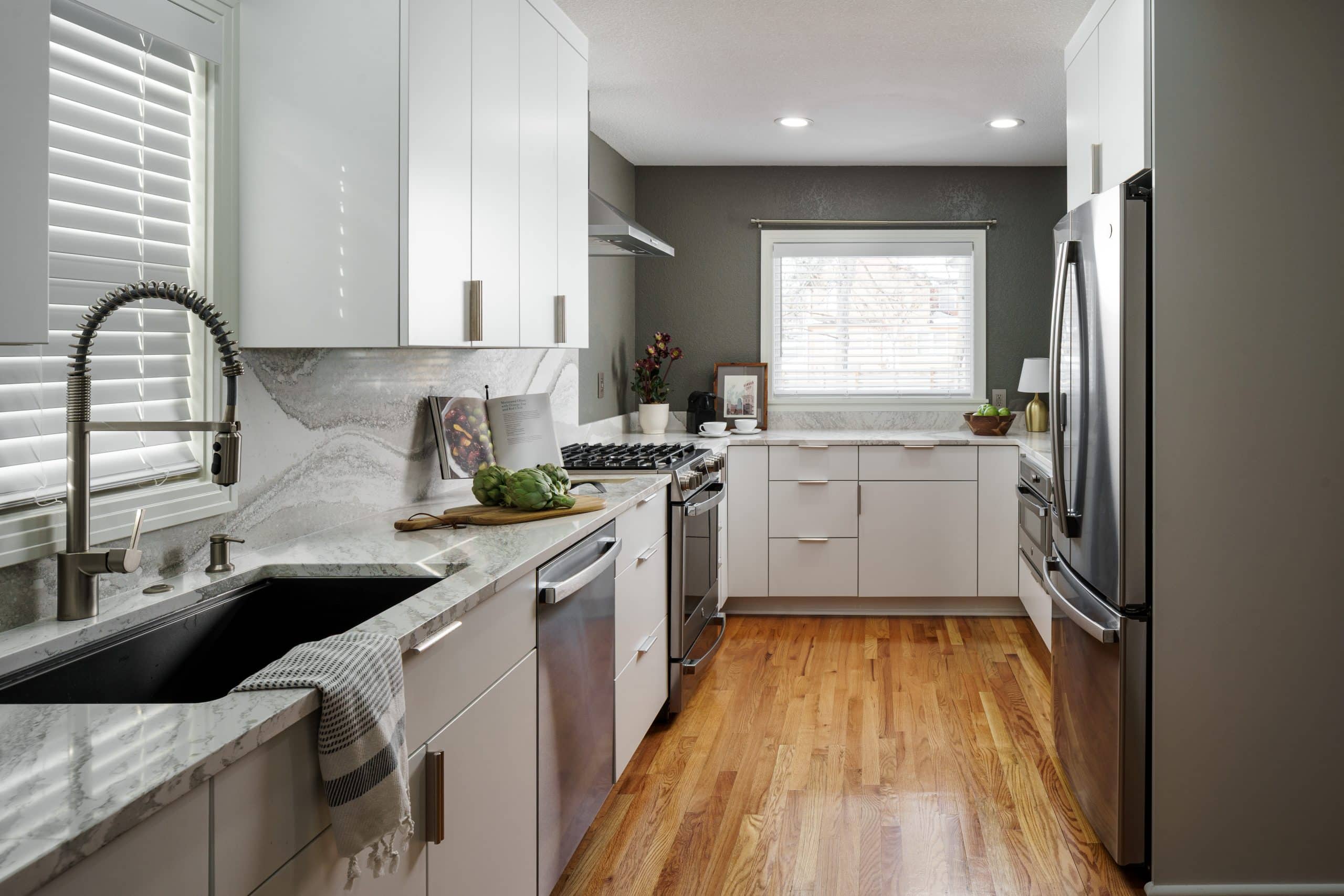 A wide view of a newly remodeled sleek, modern luxury kitchen featuring white flat-panel cabinets, light-colored quartz countertops, and a matching full-height quartz slab backsplash. A black sink with a gooseneck faucet is visible on the left, and stainless steel appliances are integrated throughout. The kitchen has warm wood flooring and a window with blinds at the far end