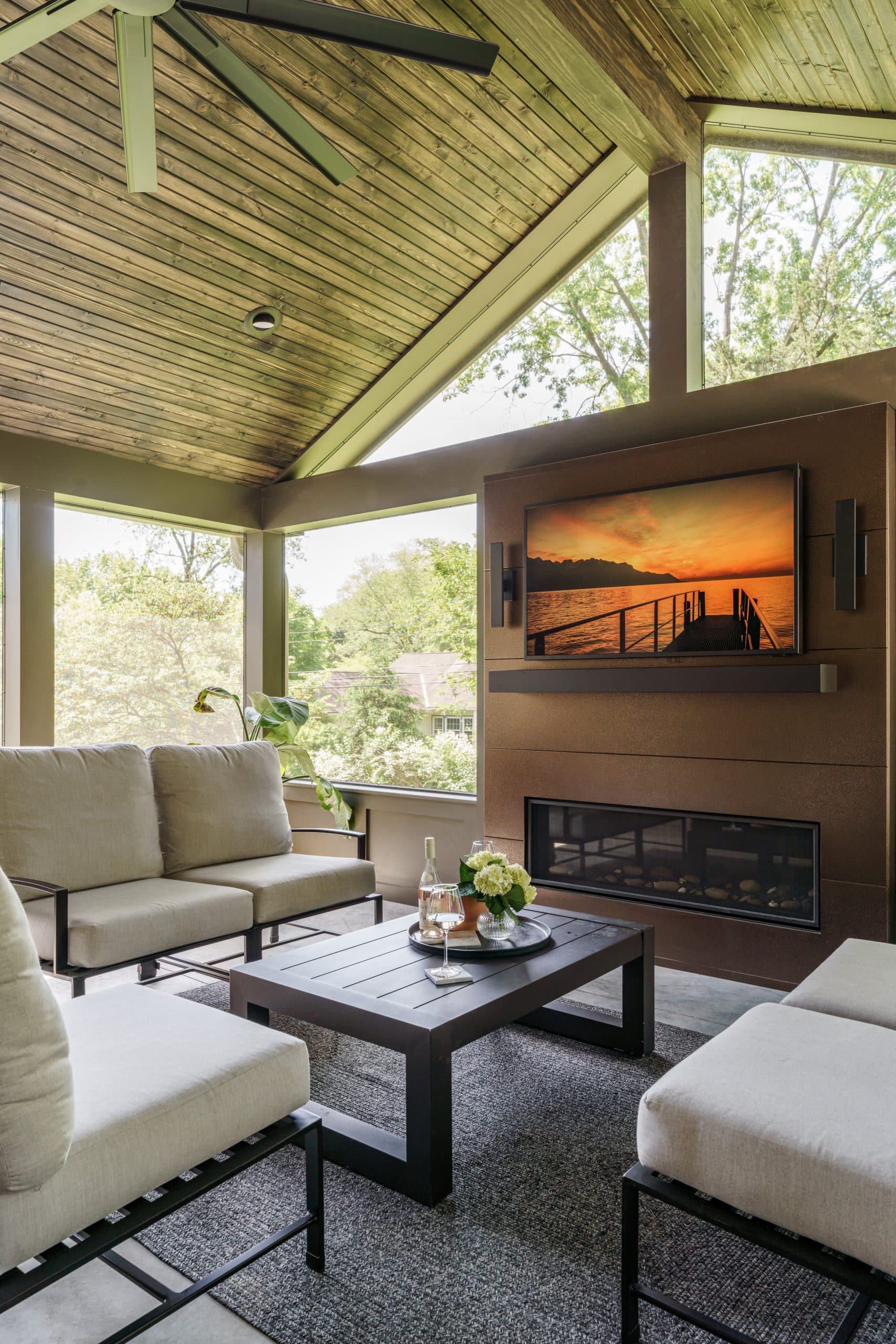 Luxurious screened-in porch addition in a Prairie Village home, featuring contemporary outdoor furniture, a linear gas fireplace, a wall-mounted television, and a warm wooden ceiling, designed for comfortable year-round use
