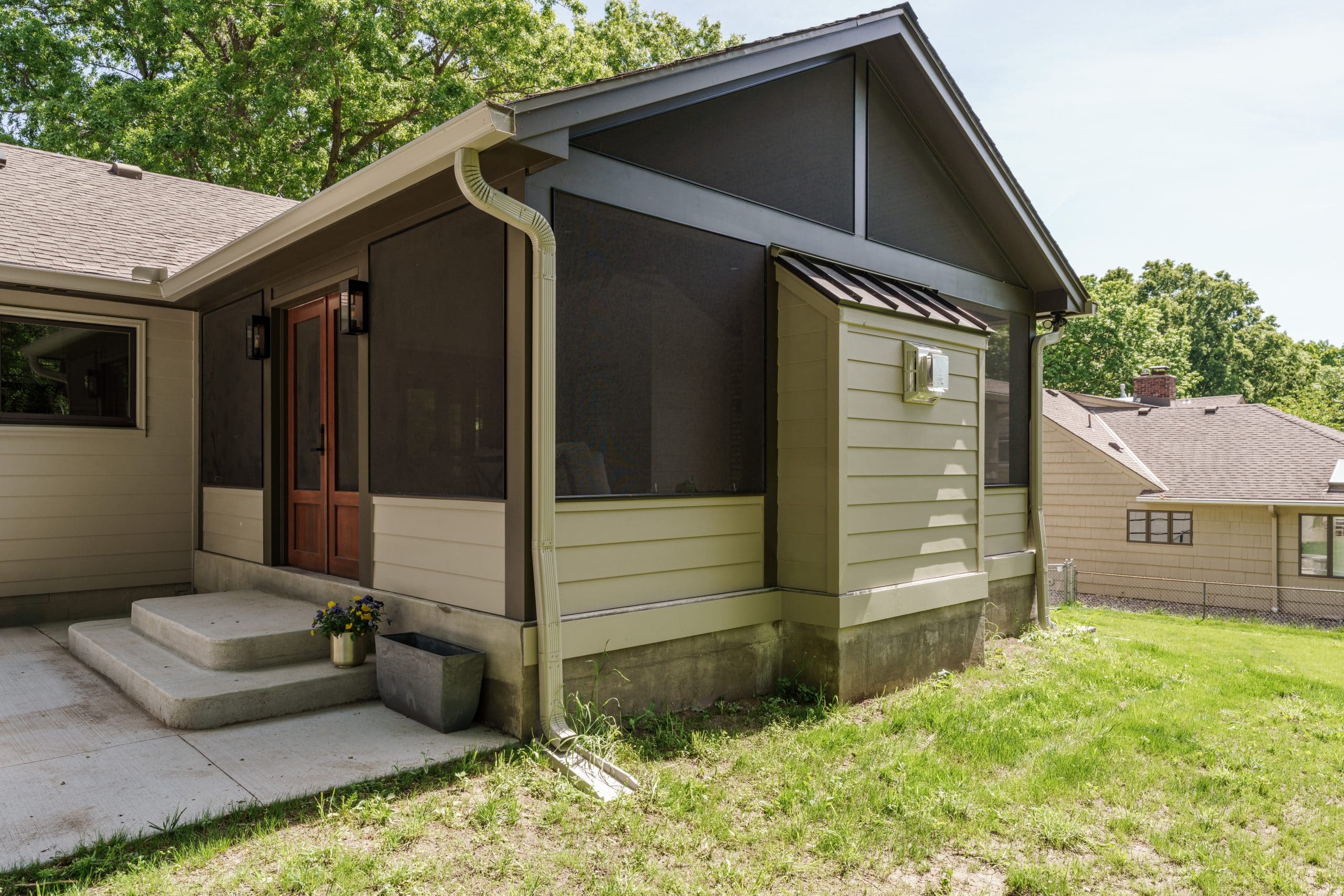Outdoor view of a modern screened-in porch with light-colored siding, a vaulted roofline, and dark brown screens, blending into the surrounding backyard.