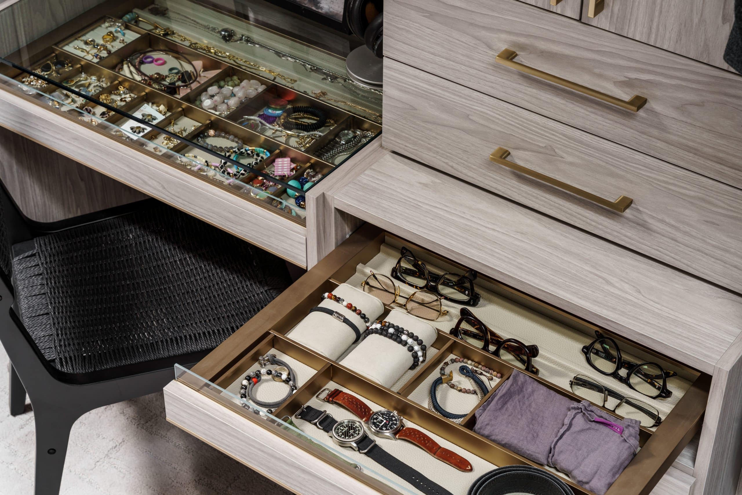 Detailed close-up of a custom-designed drawer for a walk-in closet. The image shows a sleek glass top for viewing jewelry, watches, and eyeglasses, with organized compartments. This highlights the high-end, bespoke storage solutions and modern gold hardware used in the Overland Park remodel