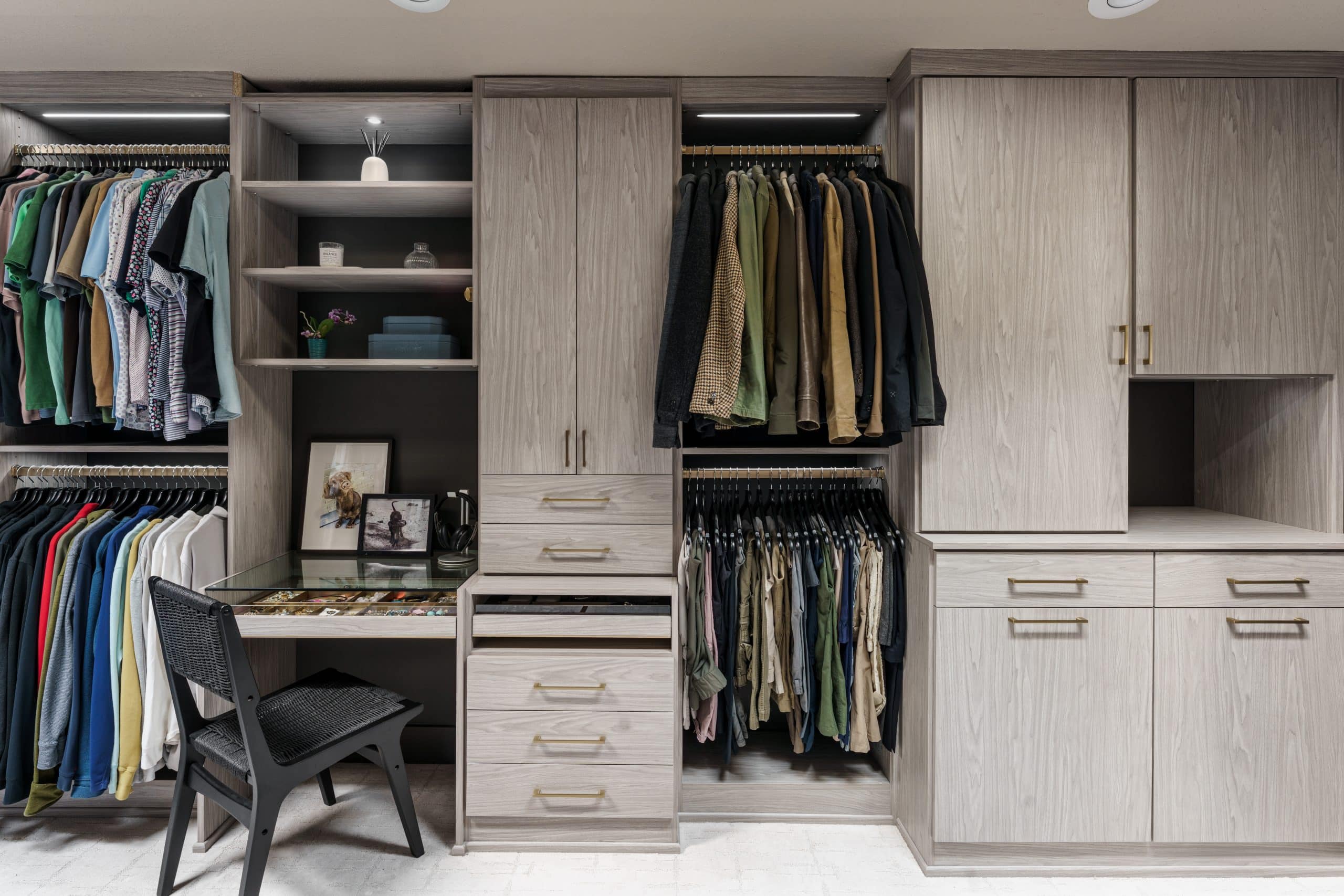 This image captures the incredible detail and custom functionality of this Overland Park closet transformation. The photo highlights the dedicated vanity and accessory area, designed for a seamless getting-ready routine. With a clear glass top, the drawers beautifully display jewelry and watches, while open shelving provides space for décor and personal items. Every element, from the custom cabinetry to the elegant gold hardware, was chosen to create a space that is both highly organized and beautifully luxurious