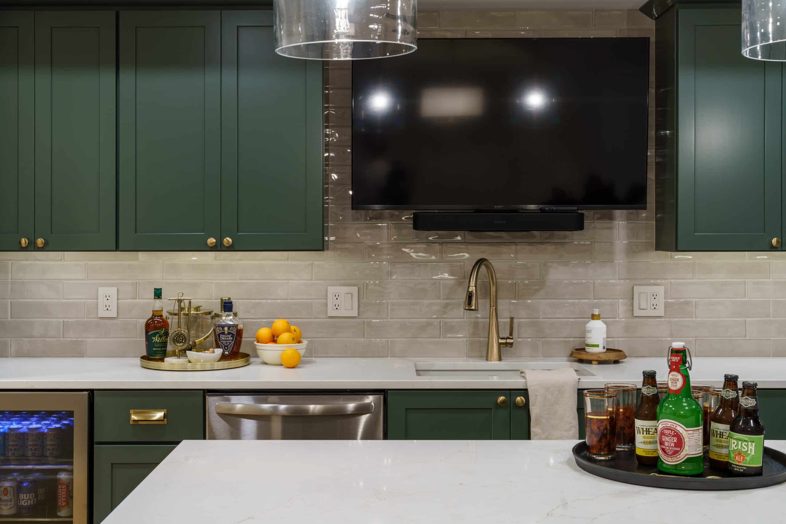 A tight, detailed interior shot of a fully equipped basement kitchen in Kansas City, showcasing a sophisticated and functional design. The image focuses on the countertops and cabinetry, which are a key part of the remodel. The custom-built upper and lower cabinets are painted a rich, deep forest green, creating a bold, modern color statement. This is contrasted with elegant gold hardware on the cabinet doors and drawers, tying into the warm palette of the overall space.
The countertops are a clean, white quartz, providing a bright and durable work surface. A subway tile backsplash in a neutral, slightly glossy finish runs the length of the counter, adding texture and a classic touch. The sink features a stylish gold-toned faucet. The kitchen is fully functional with a stainless steel dishwasher visible below the counter. A television is mounted above the sink and soundbar, perfect for watching the big game while entertaining. On the counter, various bottles and glasses are arranged, suggesting the space is ready for a party. The image also captures a small beverage fridge on the far left, emphasizing the kitchen's full hosting capabilities.