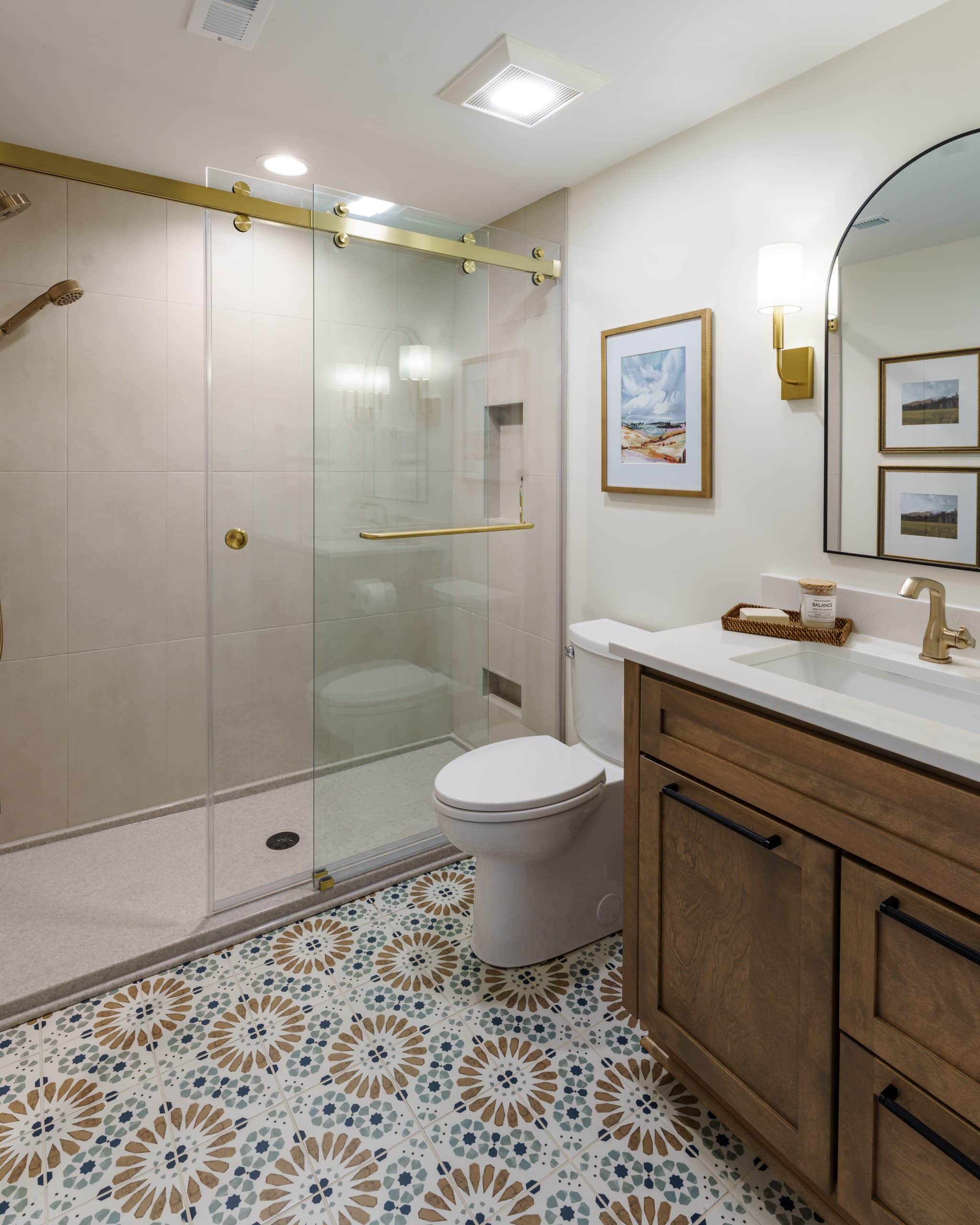 An interior shot of a newly remodeled, stylish basement bathroom, showcasing a blend of modern and eclectic design elements. The most striking feature is the patterned tile flooring with an intricate blue, brown, and white floral or starburst design, adding a vibrant and playful pop of color to the space.
The bathroom features a spacious walk-in shower with a clear glass sliding door and elegant gold-toned hardware, including the showerhead and handle. The shower walls are tiled with large, light-colored square tiles, creating a clean and seamless look.
On the right, a custom-built wood vanity with a white countertop and dark pulls provides a warm contrast to the other finishes. A large arched mirror with a thin black frame hangs above the vanity, reflecting light and making the space feel more open. Two modern wall sconces with gold accents flank the mirror, providing stylish and functional lighting. The overall design is a perfect example of how a small basement bathroom can be transformed into a bright, luxurious, and unique space.