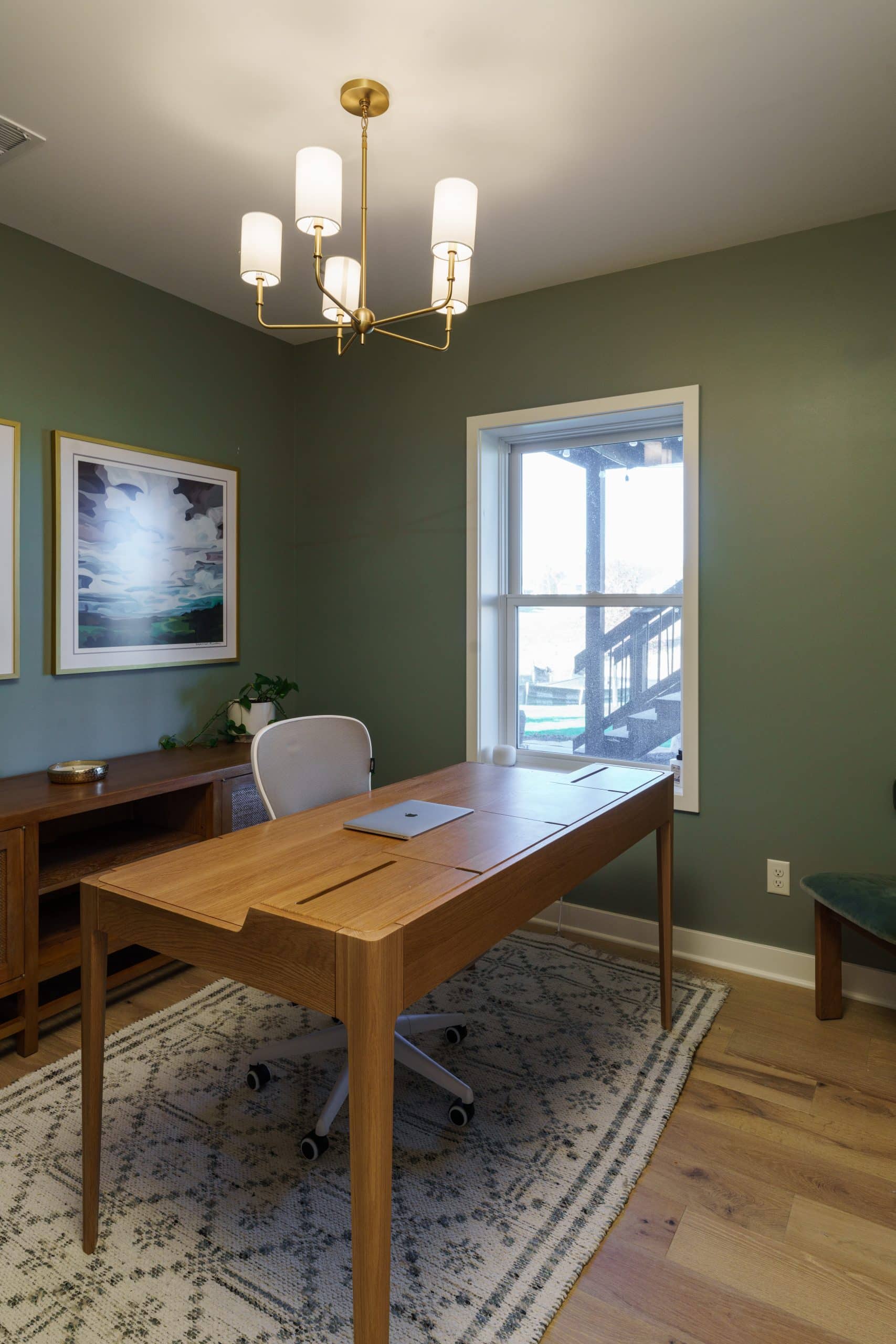 An interior shot of a newly remodeled, sophisticated home office in a basement, designed with a focus on natural light and a calming aesthetic. The walls are painted a beautiful, deep sage green, creating a peaceful and focused atmosphere. A modern, light wood desk with clean lines sits in the center of the room, positioned to take advantage of the natural light from a large window.
The room is decorated with a framed piece of art on the left wall, adding a touch of personality. The flooring is a light wood plank, which complements the warm wood tones of the desk and a low credenza on the left side of the room. A patterned area rug in neutral tones grounds the space, adding texture and warmth. A gold-toned modern chandelier with white glass shades hangs from the ceiling, providing both style and ample lighting for the workspace. This office proves that a basement can be a bright, functional, and inviting retreat.