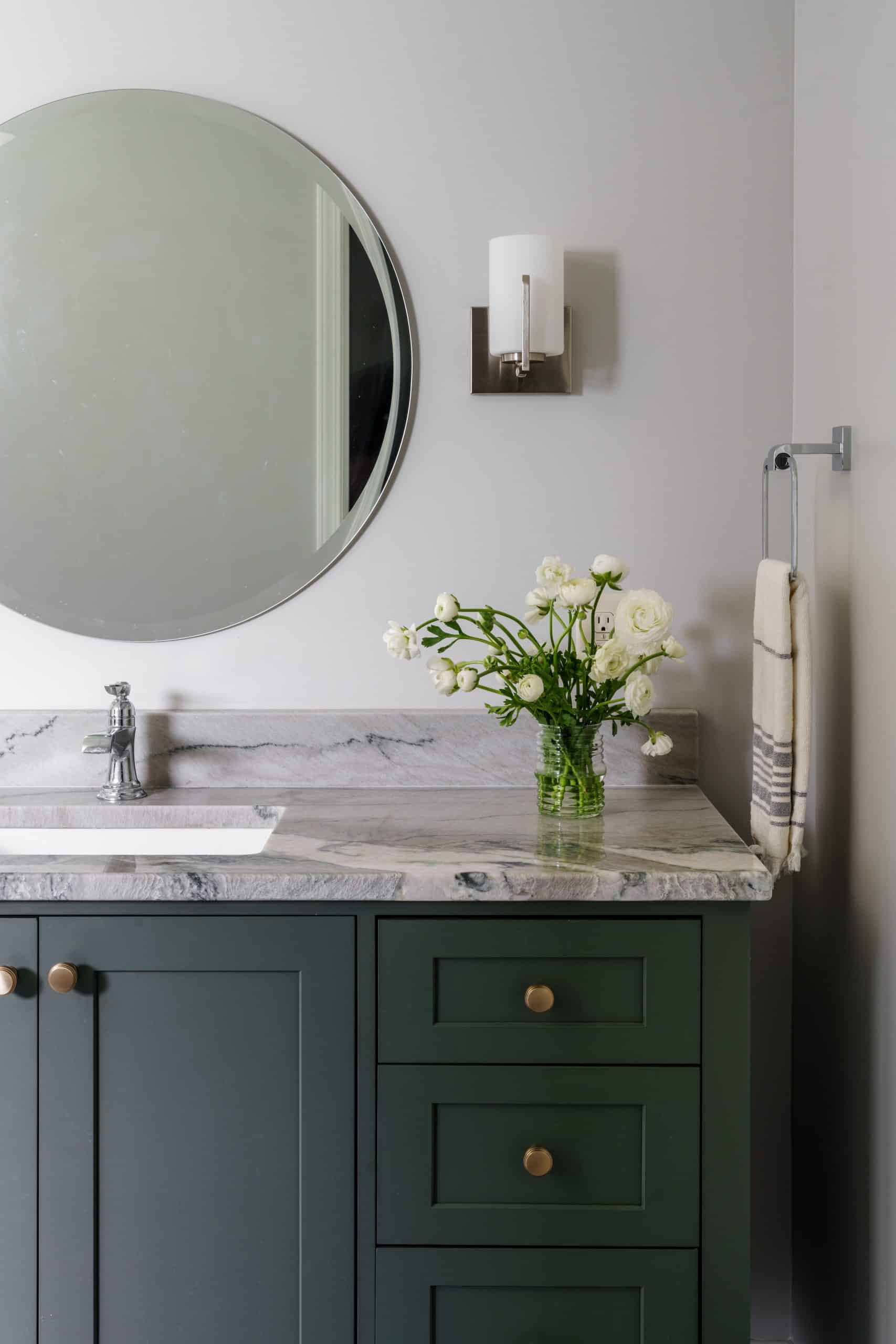 Close-up of upscale bathroom vanity detail: green shaker cabinetry, gray veined natural stone countertop, brushed brass hardware, and round mirror in a Kansas City remodel.