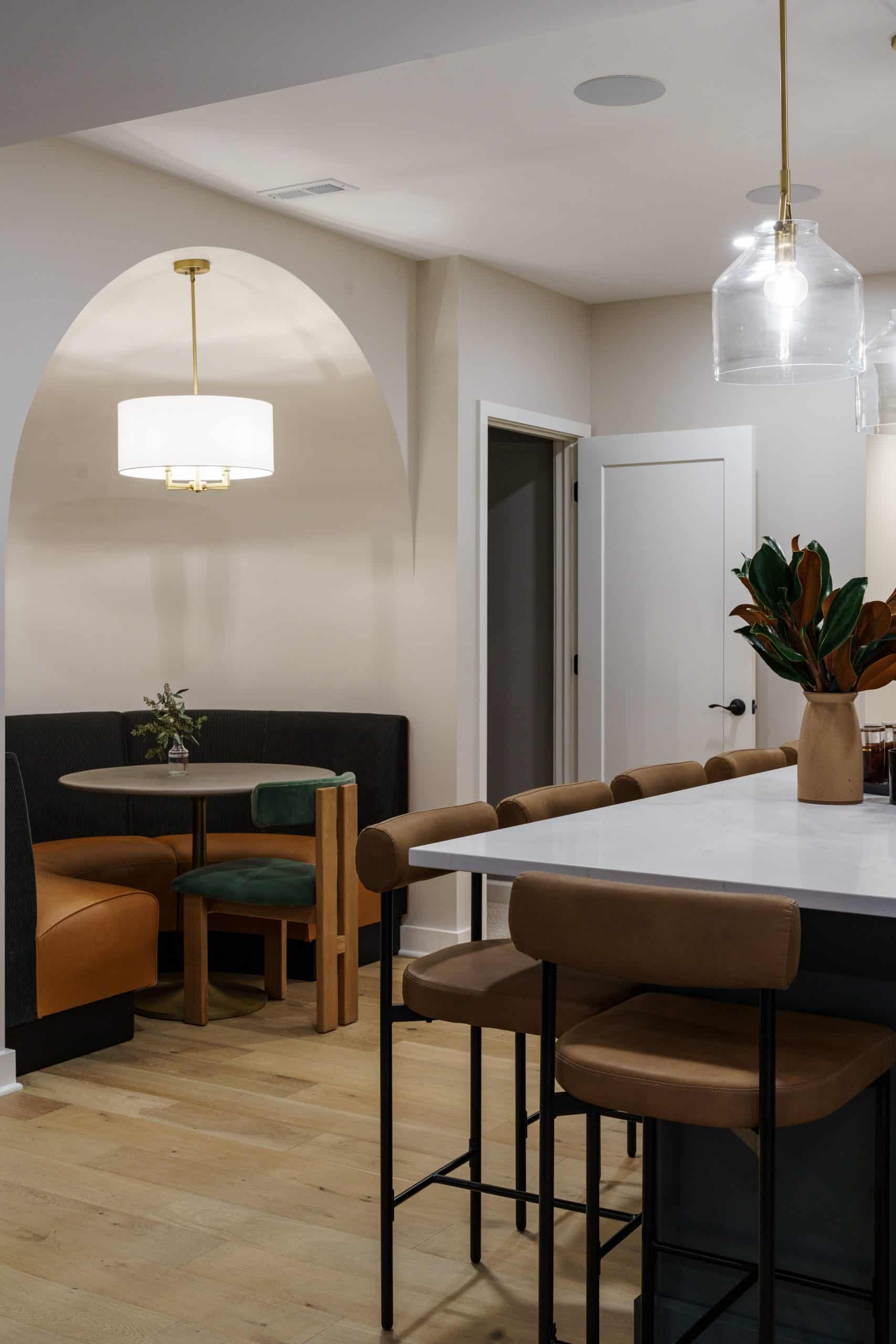 An interior detail shot of a newly remodeled basement in Kansas City, showcasing a modern and cozy dining nook. The space features a custom built-in banquette with quilted black upholstery on the back and tan leather seating, creating a stylish and inviting breakfast-nook-like area. The banquette is tucked beneath a beautifully crafted archway, which adds architectural interest and a sense of separation from the main living area.
A small round table sits in the center of the banquette, with a single green velvet chair on the opposite side, offering additional seating. A gold-toned pendant light with a white drum shade hangs above the table, providing warm and focused lighting. In the foreground, a large kitchen island with a white countertop is visible, along with two modern bar stools featuring black metal frames and tan leather seats, tying the aesthetic of the two spaces together. The flooring is a light wood plank, contributing to the overall bright and welcoming feel. This area provides a perfect, intimate spot for meals, coffee, or a quiet conversation.