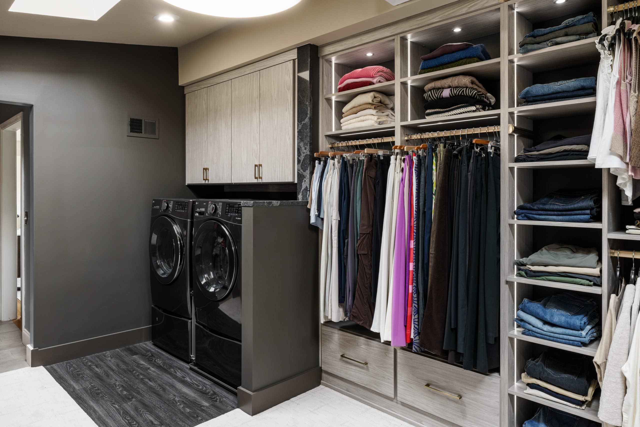Image showing the integrated laundry area within a luxury walk-in closet in Overland Park. The black washer and dryer are seamlessly installed with a custom built-in section, complemented by dark gray walls, a small section of black-patterned LVT flooring, and light-toned custom cabinetry for a modern, functional space